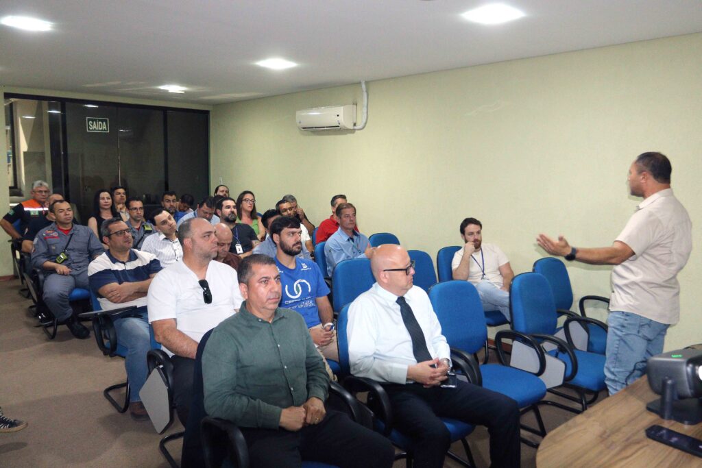 A imagem mostra uma reunião ou palestra corporativa em uma sala de conferências. Um homem está em pé à direita, falando para um grupo de pessoas sentadas em cadeiras azuis, que o observam atentamente. Os participantes parecem ser profissionais de diferentes áreas — alguns usam roupas sociais, outros uniformes ou camisetas de empresa. No fundo, há uma porta de vidro com a placa “SAÍDA” e um ar-condicionado na parede.