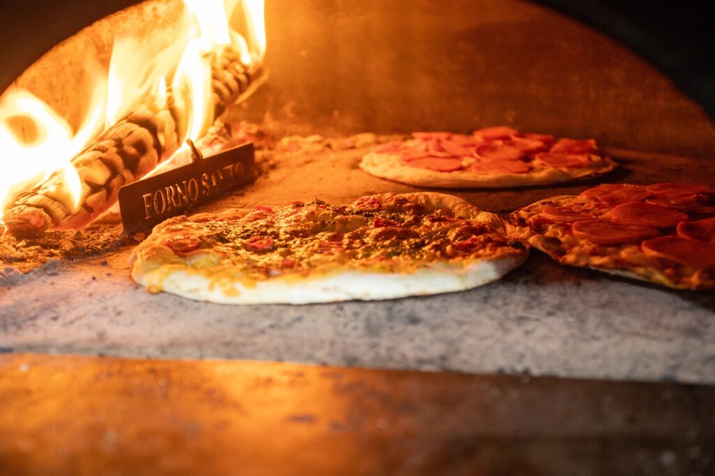 Pizza dentro de forno a lenha, sendo assada sob chama viva. Cena geral: interior de um forno a lenha com várias pizzas apoiadas diretamente na pedra refratária. Chama intensa à esquerda ilumina tudo com tons laranja e amarelo. Pizzas: massas finas, bordas levemente douradas; uma no primeiro plano com molho verde/ervas e pedacinhos de tomate, outras ao fundo com fatias de pepperoni ou salame. Algumas bordas já mostram pequenas bolhas e pontos tostados. Forno e fogo: tronco em brasa e chamas altas à esquerda; placa metálica próxima à lenha com escrita (parece “FORNO SANTO”). A pedra do forno tem cinzas e marcas escuras típicas do uso. Iluminação e textura: luz quente realçando a crocância do empanado e o brilho do queijo; textura da massa e pequenos respingos de molho visíveis em close. Atmosfera: sensação rústica e apetitosa — típico de pizzaria artesanal, preparo rápido em forno bem quente.