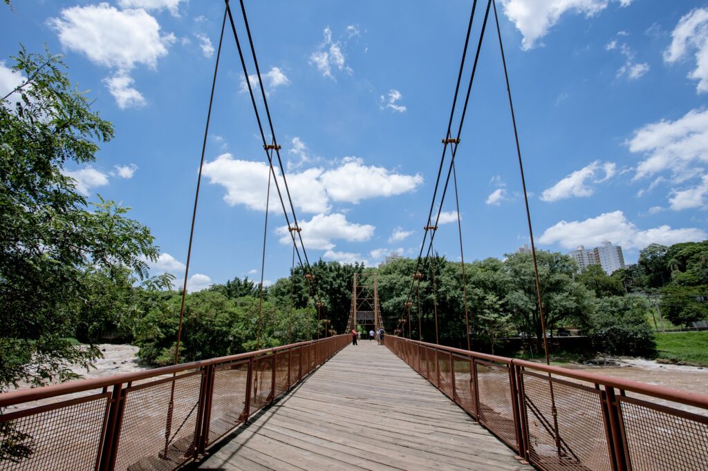 Uma ponte de suspensão de madeira se estende sobre um rio lamacento, cercada por uma vegetação exuberante e prédios altos sob um céu azul brilhante com nuvens. A imagem retrata uma perspectiva em primeira pessoa de uma ponte de suspensão de madeira vista do próprio tabuleiro. A ponte se estende para longe do observador, sua superfície de madeira composta por tábuas longas e desgastadas. Corrimãos de metal enferrujado correm ao longo de ambos os lados da ponte, protegidos por uma malha fina e em forma de diamante, que também parece enferrujada, com algumas seções mostrando um tom mais avermelhado. Vários cabos pretos verticais e grossos descem do céu, presos a suportes nas laterais do tabuleiro, contribuindo para a estrutura de suspensão da ponte. Estes cabos são sustentados por uma estrutura mais alta no meio da ponte, onde são visíveis algumas pessoas caminhando. Ao longe, no meio da ponte, há três pessoas em pé, parecendo desfrutar da vista. À esquerda da ponte, uma árvore frondosa com folhas verdes escuras se inclina sobre a cena. À direita, a paisagem é semelhante, com mais folhagem verde exuberante. Abaixo da ponte, um rio de águas turvas e marrons corre rapidamente, sugerindo uma correnteza forte. O cenário atrás da ponte é composto por uma densa folhagem de árvores verdes que cercam o rio. Mais atrás, no lado direito da imagem, vários edifícios altos e brancos aparecem acima das árvores, indicando a proximidade de uma área urbana. O céu acima é azul brilhante, pontilhado por nuvens brancas e fofas que adicionam um toque de serenidade à cena. A atmosfera geral da imagem é de um dia agradável e ensolarado, com um toque de aventura devido à estrutura imponente da ponte e ao rio turbulento abaixo. O humor é de tranquilidade e exploração, convidando o espectador a imaginar-se percorrendo a ponte e apreciando a paisagem. A combinação do céu azul, da vegetação verde e da ponte de madeira cria uma sensação de harmonia entre a natureza e a engenharia humana.