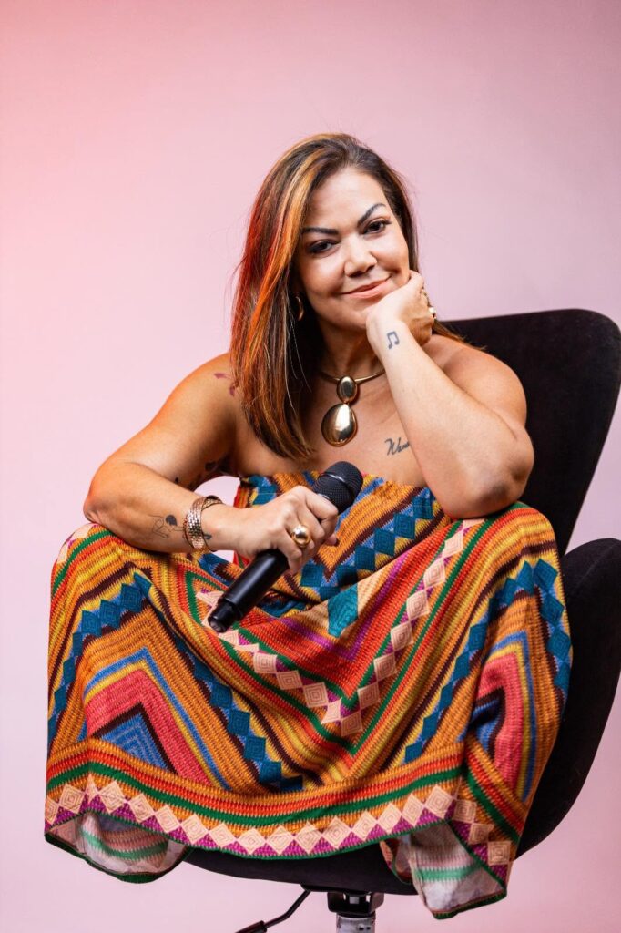 A imagem mostra um retrato em plano médio de uma mulher sorridente, sentada de forma descontraída em uma poltrona preta sobre um fundo rosa suave com iluminação aquecida. Posição e Postura: Ela está sentada de pernas cruzadas na poltrona, apoiando o queixo levemente sobre o punho esquerdo enquanto olha diretamente para a câmera com uma expressão amigável. Vestuário e Acessórios: Usa um vestido longo sem alças com estampas geométricas e étnicas bastante coloridas (tons de laranja, azul, amarelo, verde e vermelho). Complementa o visual com um colar dourado marcante em formato de gota, pulseira e anéis dourados. Objeto na Mão: Na mão direita, repousada sobre o colo, ela segura um microfone preto. Tatuagens e Cabelo: Possui pequenas tatuagens visíveis no punho (uma nota musical), no antebraço e no tórax. Seu cabelo é castanho na altura dos ombros, com mechas mais claras em tom mel.