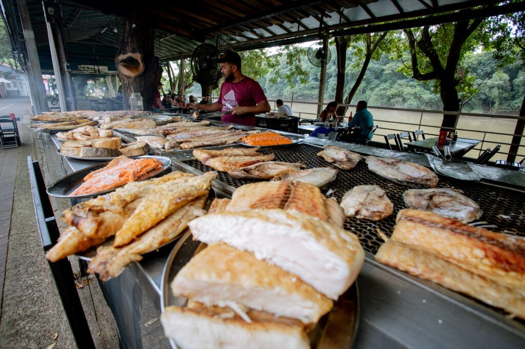 A imagem mostra um restaurante ao ar livre, às margens de um rio, especializado em peixes preparados na brasa.
Em primeiro plano, há uma grande variedade de peixes assados e grelhados, dispostos em bandejas e sobre uma extensa grelha. Os peixes apresentam coloração dourada e algumas peças têm a carne alaranjada, criando um forte destaque visual.
Ao centro, um funcionário do restaurante trabalha junto à churrasqueira, enquanto, ao fundo, alguns clientes estão sentados às mesas. O espaço é coberto por uma estrutura simples, com ventiladores e elementos rústicos, incluindo uma grande árvore integrada ao ambiente.
Do lado direito, é possível observar a vegetação às margens do rio, reforçando o caráter natural e turístico do local. A cena transmite uma atmosfera rústica, tradicional e gastronômica, destacando o preparo de peixes na brasa como uma das principais atrações do restaurante.