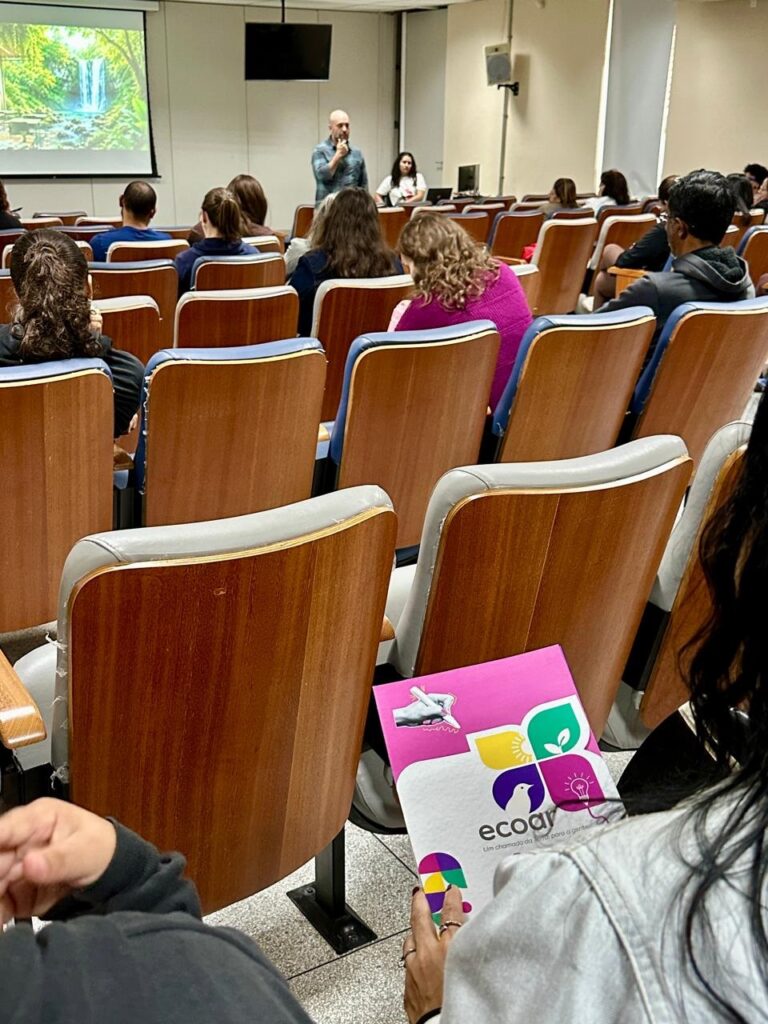 A imagem mostra um evento, possivelmente uma palestra ou conferência, em um auditório. Destaque: Em primeiro plano, uma pessoa segura um material informativo (como um panfleto) da iniciativa "ecoar", com a frase "Um chamado da terra para a gente". O logotipo é bastante colorido e contém ícones que remetem a natureza e ideias (sol, folha, pássaro e lâmpada). Palestra: Ao fundo, um homem está falando no palco, em frente a uma tela que projeta a imagem de uma cachoeira em meio a uma floresta. Ambiente: O público está acomodado em poltronas de madeira com assentos estofados, prestando atenção à apresentação. O contexto visual sugere um evento voltado para a sustentabilidade ou educação ecológica.