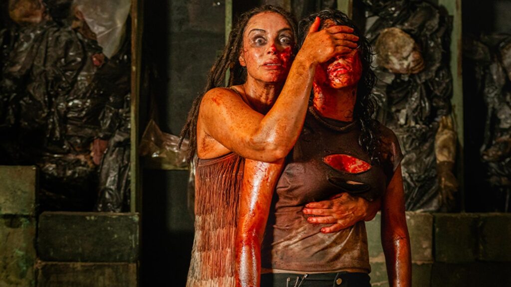 A imagem mostra uma cena de terror com duas mulheres cobertas de sangue e sujeira. A mulher ao fundo, com os olhos arregalados, segura a cabeça da outra, que tem o rosto parcialmente coberto pela mão e veste uma camiseta escura rasgada. Ao redor delas há sacos plásticos pretos e um ambiente escuro, com iluminação avermelhada e aparência abandonada.