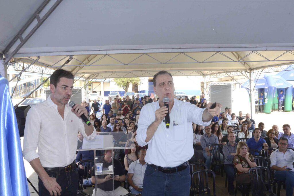 A imagem mostra uma cerimônia ou evento público realizado em um espaço coberto, com uma grande plateia sentada ao fundo. Em primeiro plano, há três homens diante de um púlpito de acrílico: dois usam camisas brancas e um veste uma camisa listrada clara. Um dos homens segura um microfone e parece falar ao público, enquanto os outros o acompanham. À esquerda, há um painel azul com o brasão e a identidade visual da Prefeitura de Piracicaba. O local possui estrutura metálica, cobertura clara, cadeiras e decoração em tons de azul e verde. A imagem está parcialmente superexposta e corta parte das pessoas nas bordas.