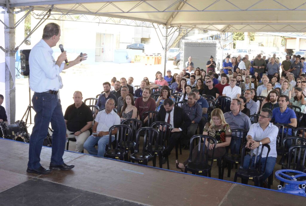 A imagem mostra um homem de camisa branca falando ao microfone diante de uma grande plateia sentada. Ele está em um palco, ao lado de equipamentos de som, enquanto o público acompanha atentamente a apresentação. O evento acontece em um espaço coberto, com estrutura metálica e iluminação natural intensa. Ao fundo, aparecem prédios, árvores e veículos, indicando um ambiente aberto ou parcialmente externo. A fotografia está bastante clara e superexposta em algumas áreas.