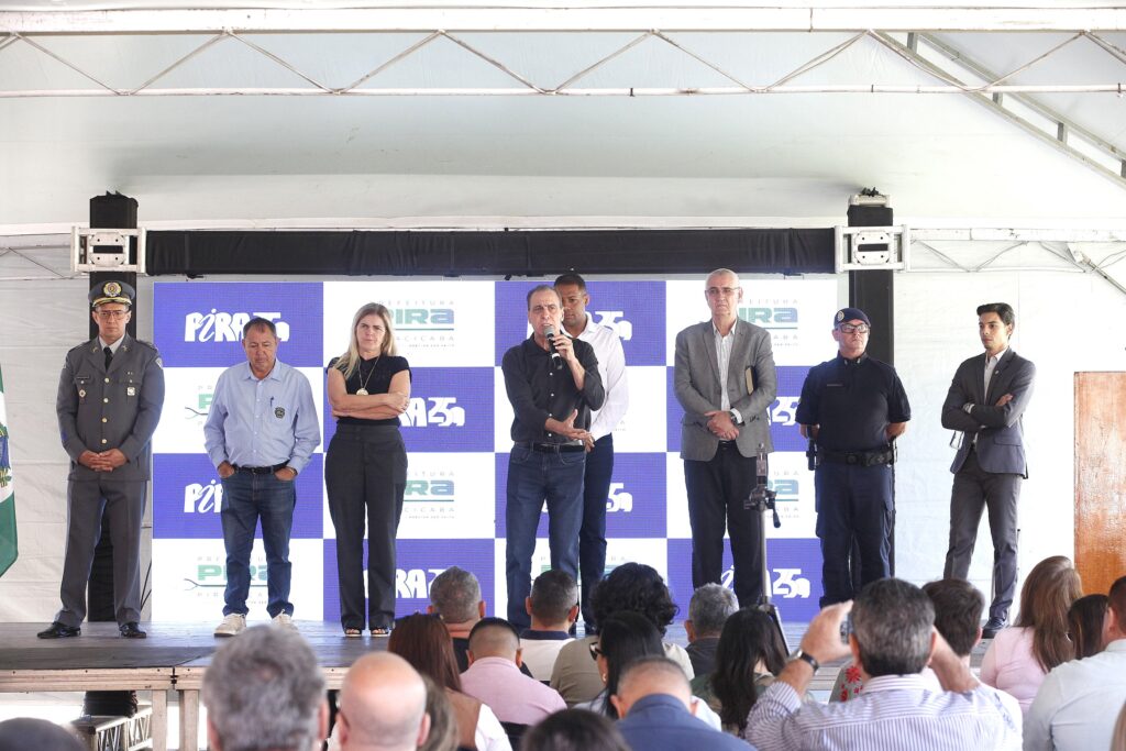 A imagem mostra um evento oficial ou uma coletiva de imprensa ocorrendo em um palco coberto. No centro, um homem de camisa preta está falando ao microfone. Ele está cercado por várias outras pessoas, formando uma linha: À esquerda do orador: Uma mulher de blusa preta com os braços cruzados, um homem de camisa azul clara e jeans, e um oficial da Polícia Militar em uniforme de gala. À direita do orador: Um homem alto de camisa branca, um homem de terno cinza, um guarda municipal (ou autoridade de segurança) fardado e um homem jovem de terno escuro. O fundo é um painel azul e branco com a marca "Pira" (referência à Prefeitura de Piracicaba). Diante do palco, há uma plateia assistindo à apresentação, com várias pessoas de costas para a câmera.