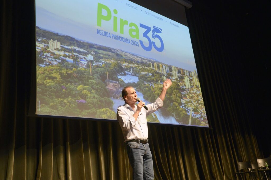 Um homem de meia-idade faz uma apresentação dinâmica em um auditório, gesticulando enquanto fala diante de um grande telão que projeta o logotipo "Pira35" e imagens aéreas de uma cidade. A composição da cena é centralizada e focada no orador, que ocupa o primeiro plano, criando um contraste direto entre sua figura ativa e a imagem estática projetada ao fundo. O homem está posicionado ligeiramente à direita do centro, com um braço levantado em um gesto enfático, sugerindo um momento de discurso persuasivo ou explicativo. A moldura do palco, com suas cortinas escuras nas laterais, direciona o olhar do espectador para o centro, onde a luz incide sobre o apresentador, isolando-o contra o brilho suave da tela de projeção. O sujeito principal é um homem de meia-idade, com cabelos curtos e castanhos, vestindo uma camisa social clara de mangas compridas, levemente dobradas, e calças jeans escuras. Sua expressão é de concentração e envolvimento, com a boca aberta como se estivesse articulando uma palavra importante. Ele segura um microfone com a mão direita, enquanto sua mão esquerda está erguida, complementando sua postura comunicativa. A sua vestimenta sugere um ambiente de negócios ou evento público, transmitindo uma imagem de formalidade profissional, porém acessível. A imagem é uma fotografia de evento com iluminação técnica controlada. A execução técnica privilegia a clareza do orador, mantendo o fundo com a projeção visível, mas levemente fora de foco em relação ao homem em destaque. O uso da luz é estratégico: o sujeito é iluminado por uma fonte frontal que o destaca das cortinas escuras atrás, enquanto o brilho do projetor serve como luz de fundo, criando um contorno suave ao redor do orador. O estilo é puramente documental, capturando a essência de uma apresentação pública. O ambiente é um auditório com pouca iluminação, o que ajuda a enfatizar a projeção na tela e o orador. As cortinas pesadas e escuras nas laterais conferem um tom sóbrio e institucional ao local. A atmosfera é de conferência, palestra ou seminário, com uma sensação de seriedade e foco no tema abordado, que parece ser um projeto de desenvolvimento urbano ou agenda estratégica intitulada "Pira35". A iluminação focalizada cria um ambiente intimista e concentrado, garantindo que a atenção do público esteja totalmente voltada para o orador e a mensagem visual que ele apresenta.
