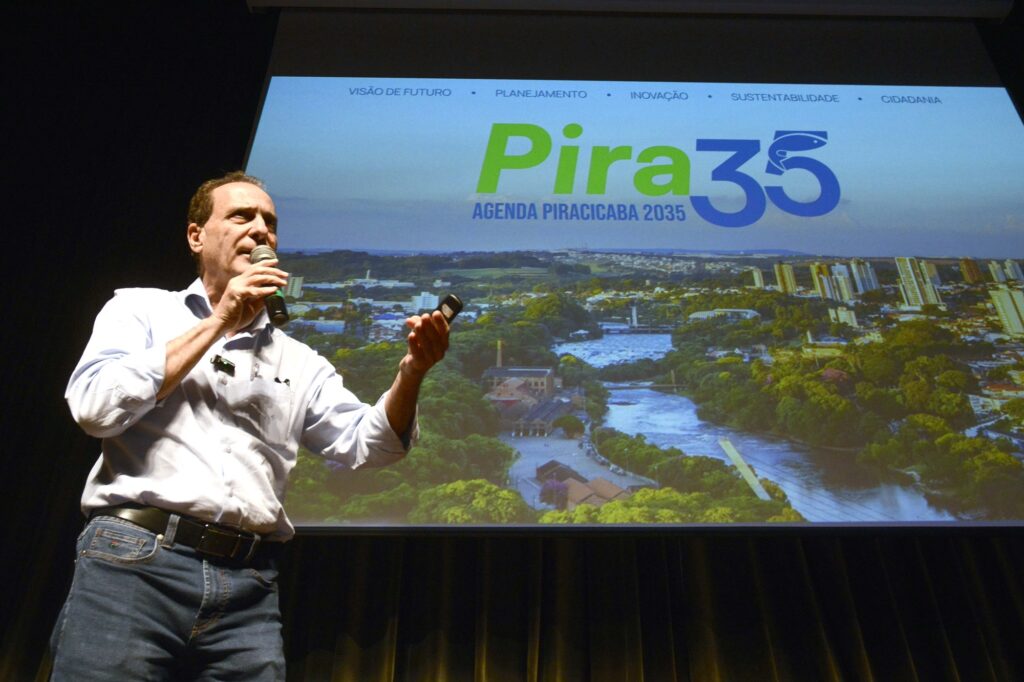A imagem mostra um homem fazendo uma apresentação em um evento, segurando um microfone em uma das mãos e um controle remoto de apresentação na outra. Ao fundo, há um grande telão exibindo a identidade visual “Pira 35 – Agenda Piracicaba 2035”, sobreposta a uma imagem panorâmica da cidade de Piracicaba, com destaque para o rio, áreas verdes e edifícios urbanos. Na parte superior do telão aparecem palavras-chave que parecem orientar o projeto: “Visão de Futuro”, “Planejamento”, “Inovação”, “Sustentabilidade” e “Cidadania”. A composição transmite um contexto de lançamento ou apresentação institucional, relacionado ao planejamento estratégico e ao futuro do município.