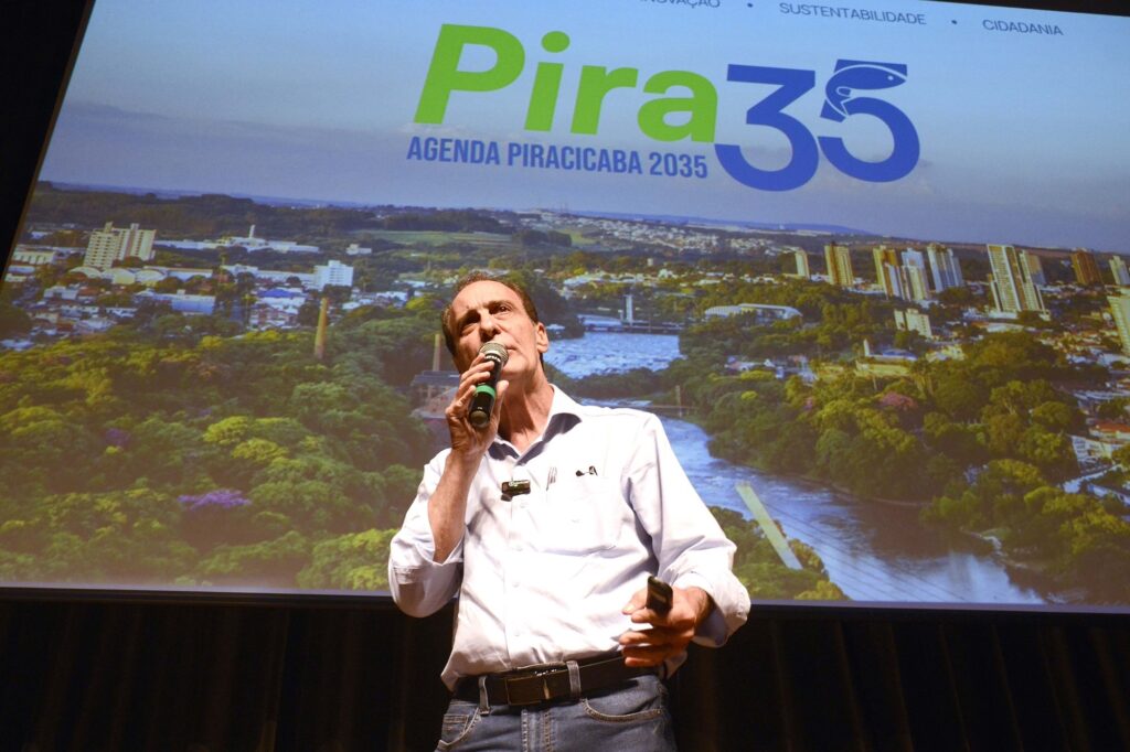 A imagem mostra um homem de camisa branca falando ao microfone em um palco ou auditório. Ele segura um controle na outra mão e parece estar fazendo uma apresentação. Ao fundo, há uma grande projeção com uma vista aérea de Piracicaba, incluindo áreas urbanas, vegetação e um rio. No topo, aparece o texto “Piracicaba 35 — Agenda Piracicaba 2035”, acompanhado de palavras como “inovação”, “sustentabilidade” e “cidadania”.