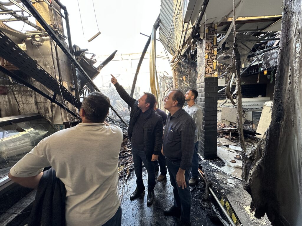 A imagem mostra quatro pessoas em meio às ruínas carbonizadas de um prédio que foi devastado por um incêndio. O cenário é marcado por vigas metálicas retorcidas, paredes enegrecidas e destroços espalhados pelo chão, transmitindo a intensidade da destruição. Uma das pessoas aponta para cima, possivelmente indicando a gravidade dos danos ou algum detalhe relevante na estrutura.
