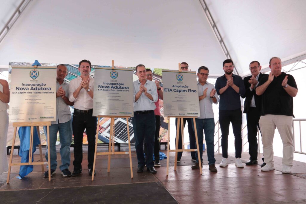 A imagem mostra um grupo de pessoas em uma cerimônia de inauguração. Elas estão em um espaço coberto, posicionadas atrás de placas ou painéis com o brasão de Piracicaba e os dizeres “Inauguração”, “Nova Adutora” e “ETA Capim Fino – Torre de TV”. Alguns participantes usam roupas sociais, enquanto outros vestem camisetas ou uniformes azuis. Ao fundo, há uma estrutura metálica e uma tela ou painel com imagens. A fotografia está parcialmente cortada, deixando bastante espaço vazio na parte superior e mostrando apenas parte do corpo das pessoas.