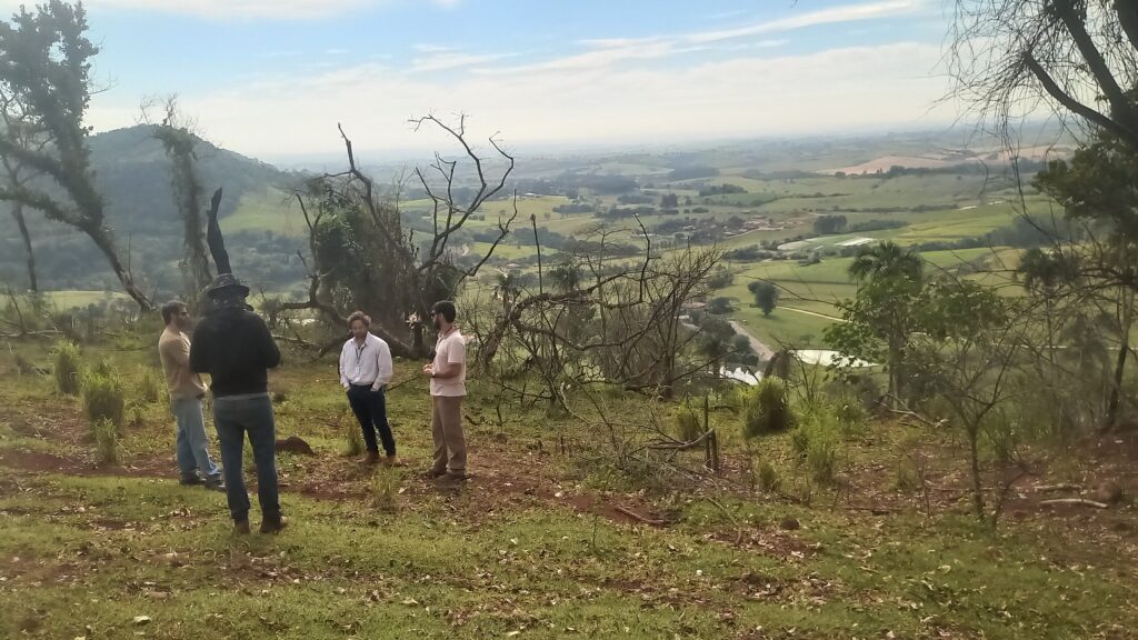 Nesta imagem, vemos quatro homens em pé em uma colina, observando uma vasta paisagem rural. Eles parecem estar em um momento de conversa ou análise, talvez avaliando o terreno ou a vista. A cena transmite uma sensação de tranquilidade, com uma vista aberta que inclui campos verdes, colinas suaves e, ao longe, o que parece ser uma área habitada ou uma propriedade rural. O dia está um pouco nublado, mas iluminado, e o local onde eles estão parece ter solo de terra batida com algumas árvores e vegetação rasteira.
