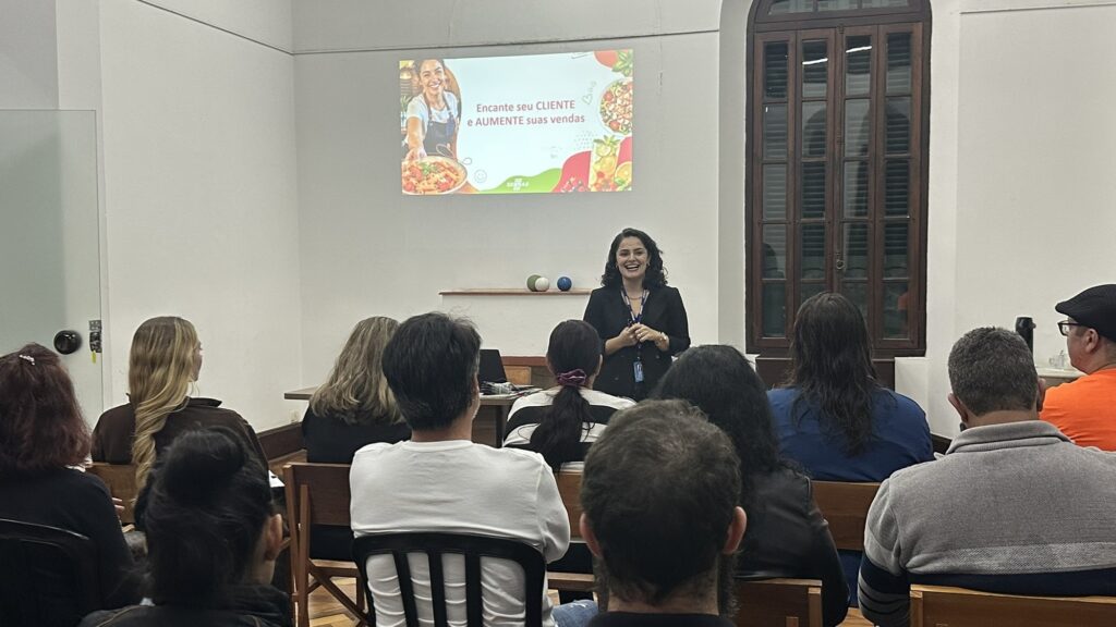 A imagem mostra uma palestra ou treinamento em uma sala. Uma mulher está em pé na frente do público, sorrindo e falando com os participantes, que estão sentados de costas para a câmera. Na parede, há uma projeção com o texto “Encante seu CLIENTE e AUMENTE suas vendas”, acompanhada de imagens de alimentos e do logotipo do Sebrae. A sala tem paredes claras, janelas de madeira e uma mesa com um notebook perto da palestrante.