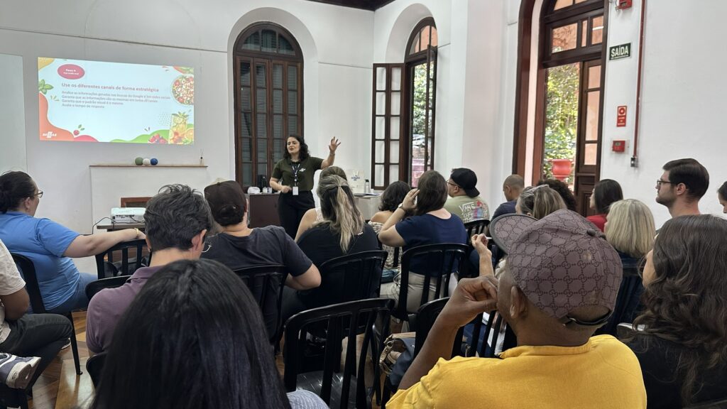 A imagem mostra uma apresentação/treinamento ministrado em uma sala ampla de estilo arquitetônico clássico. No centro do espaço, uma mulher vestindo uma camiseta verde-musgo com crachá e calça escura está em pé, gesticulando com a mão esquerda enquanto fala para a plateia. À esquerda, há uma projeção na parede com o logotipo do SEBRAE no canto inferior. O título do slide diz "Use os diferentes canais de forma estratégica" e traz pontos sobre analisar informações em buscas do Google/redes sociais, manter padrão visual e avaliar tempo de resposta. Diversas pessoas aparecem sentadas em cadeiras pretas, de costas ou de perfil para a câmera, prestando atenção à palestra. Em primeiro plano, destaca-se um homem com camiseta amarela e boné marrom. A sala possui paredes brancas altas, janelas e portas em arco com caixilhos de madeira e vidro. Há uma placa verde de "SAÍDA" visível acima de uma das portas à direita.