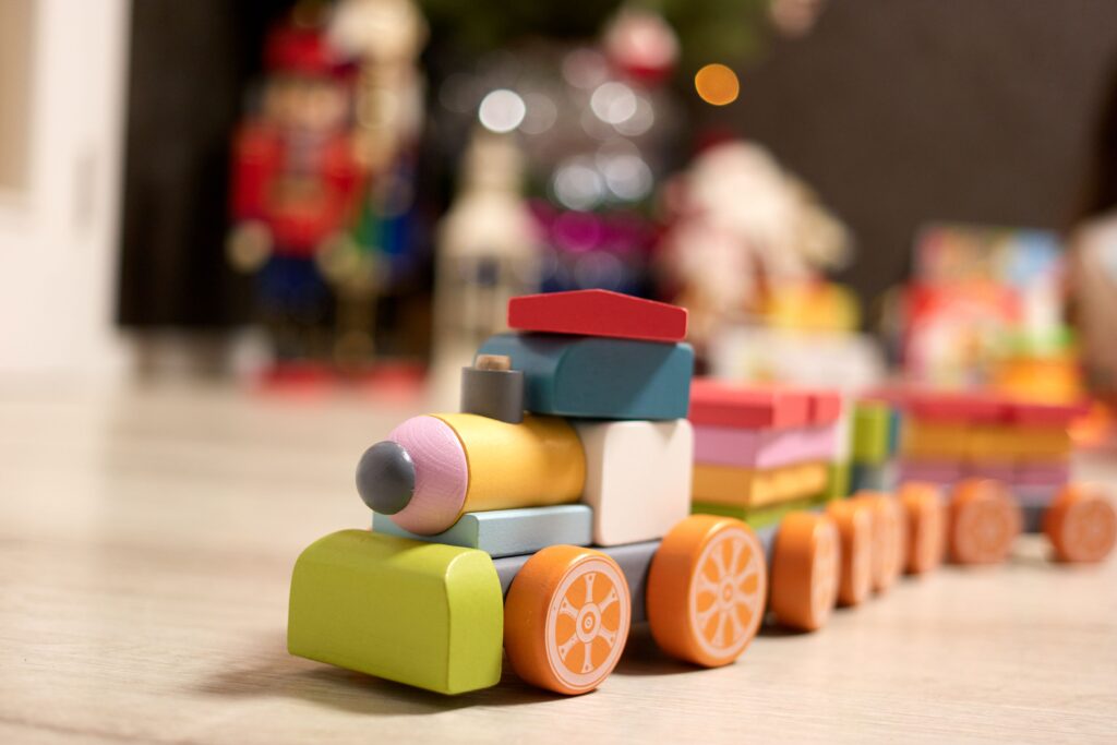 Um encantador trem de brinquedo feito de blocos de madeira coloridos repousa sobre uma superfície clara, evocando uma sensação nostálgica de diversão infantil em um ambiente doméstico festivo e acolhedor. A composição da cena é centrada no trem de madeira, posicionado em um ângulo diagonal que guia o olhar do observador da parte frontal, com seu para-choque verde, em direção aos vagões traseiros que desaparecem suavemente no fundo. O brinquedo ocupa a posição de destaque, enquanto o fundo está intencionalmente desfocado, criando um efeito de profundidade de campo que isola o objeto e enfatiza sua presença. O enquadramento é próximo e ao nível do solo, permitindo uma conexão visual íntima com o brinquedo. O sujeito principal é um trem de montar de madeira composto por formas geométricas distintas. A locomotiva possui uma frente arredondada em tons de rosa e cinza, um corpo principal em azul e verde, e uma chaminé pequena e charmosa. Os vagões subsequentes são formados por blocos empilhados de cores variadas — amarelo, rosa, verde e laranja — que conferem ao brinquedo uma aparência lúdica e artesanal. As rodas do trem são pintadas com detalhes radiais que lembram o design de laranjas fatiadas, adicionando um toque criativo e artístico ao conjunto. A execução técnica revela um foco na fotografia macro, onde a nitidez é mantida estritamente no corpo do trem, enquanto o restante da cena é dissolvido em um borrão suave. O estilo é naturalista e focado na captura de texturas, realçando o acabamento liso e fosco da madeira pintada do brinquedo. A escolha de cores vibrantes, mas com tons pastéis e terrosos, confere ao trabalho um aspecto esteticamente agradável e equilibrado, típico de brinquedos educativos de alta qualidade voltados para o desenvolvimento infantil.