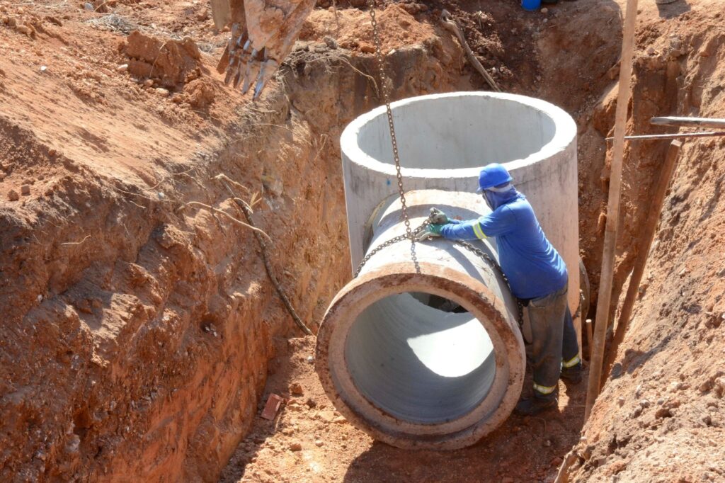 Na imagem, vemos um trabalhador de construção civil posicionando grandes tubos de drenagem de concreto dentro de uma vala profunda escavada em solo avermelhado. Ele usa capacete azul, luvas e camisa de mangas compridas, enquanto o braço de uma escavadeira com corrente auxilia na descida e alinhamento das peças cilíndricas pesadas. A cena retrata um momento crucial da instalação de infraestrutura subterrânea, como sistemas de esgoto ou drenagem pluvial, destacando a combinação de força mecânica e precisão manual necessária para garantir o encaixe correto dos componentes.