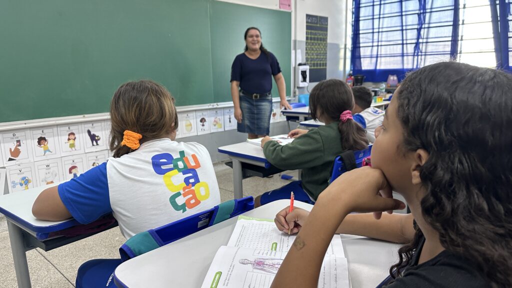 A imagem mostra uma sala de aula. Uma professora está em pé diante de um quadro verde, sorrindo, enquanto várias crianças permanecem sentadas em carteiras azuis e brancas, realizando atividades em folhas de papel. No fundo, há janelas grandes com cortinas azuis, armários e materiais escolares. Na parede abaixo do quadro, aparecem cartazes com letras do alfabeto e ilustrações. Em primeiro plano, uma aluna usa uma camiseta branca e azul com letras coloridas, e outra criança escreve com lápis em uma atividade que inclui o desenho do corpo humano.