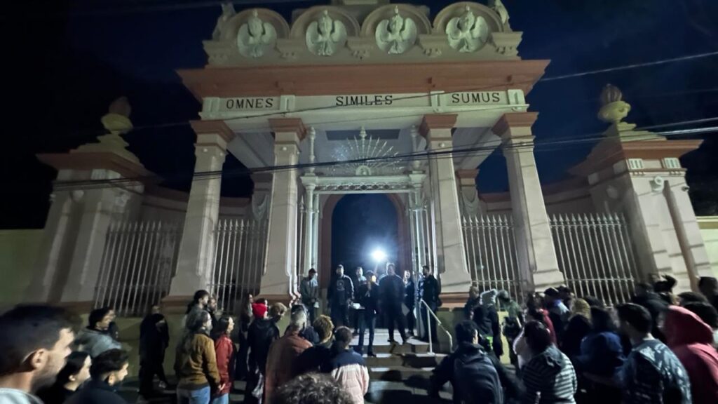 Entrada monumental de um edifício histórico à noite, com um grupo grande de pessoas reunidas na frente e nos degraus. Arquitetura: portão imponente com colunas e um pórtico decorado em tons claros (creme/rosa). No entablamento está escrito em latim “OMNES SIMILES SUMUS” (letras maiúsculas). Ornamentos circulares e esculturas discretas no topo, grade metálica alta dos dois lados e um arco de passagem central. Iluminação: cena predominantemente escura, iluminada por luzes frontais fortes (há uma fonte de luz bem intensa dentro do arco, quase em direção à câmera), criando contrastes e silhuetas; sombras longas e destaque nos rostos voltados para a entrada. Pessoas: dezenas de pessoas reunidas na calçada e sobre os degraus, algumas em pequenos grupos conversando ou observando; várias vestem casacos e capuzes (sugere clima frio). Ninguém é identificado individualmente — aparecem como participantes de uma reunião pública. Ação e atmosfera: sensação de encontro ou assembleia pública — a maioria olha para o arco/porta onde algumas pessoas estão posicionadas (possivelmente falando ou organizando algo). O clima parece sério, atento e coletivo, com foco no ponto iluminado central. Detalhes menores: corrimão central até o arco, postes e fios elétricos na frente do prédio, movimentos e aglomeração indicam evento noturno em espaço urbano.