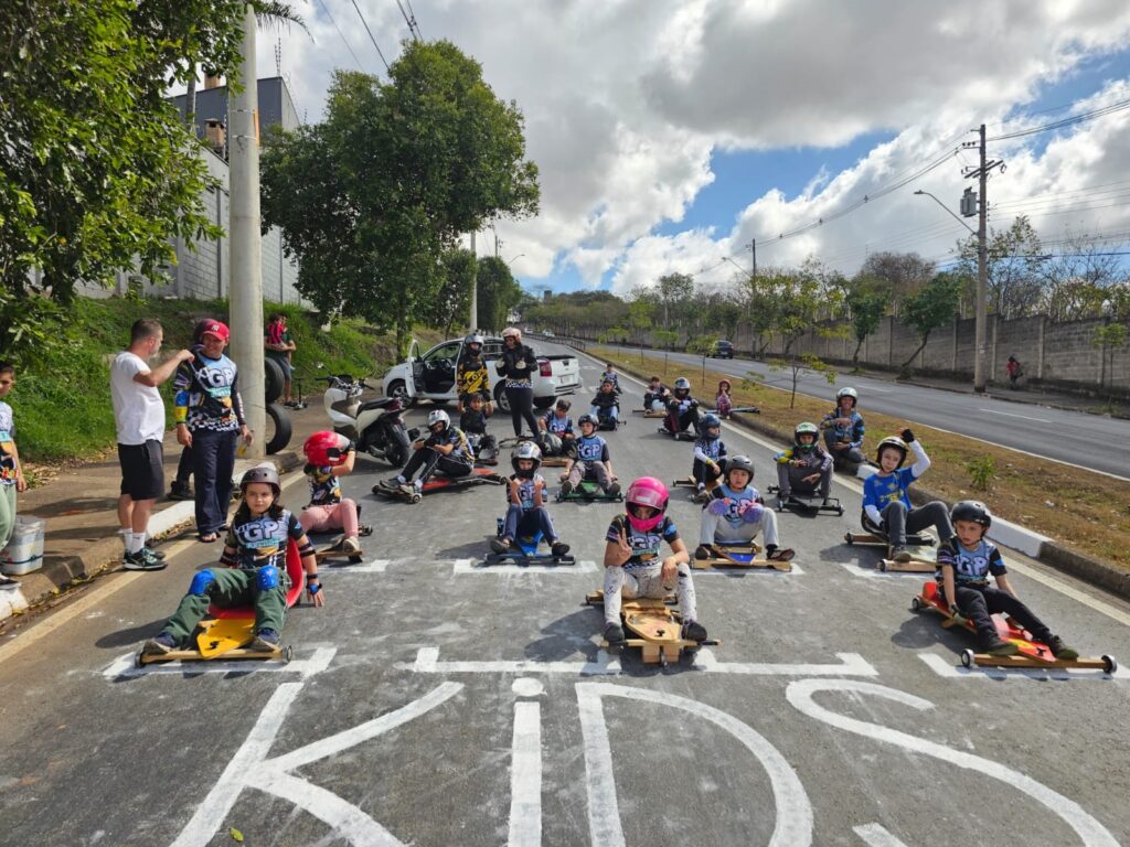 Vejo uma largada de evento de carrinhos de madeira numa via fechada ao tráfego: Grupo: cerca de 16 a 20 crianças e adolescentes sentados em carrinhos de madeira/lowriders, dispostos em filas na faixa de largada pintada no asfalto. Segurança: quase todos usam capacete; alguns também têm joelheiras e luvas. Muitos vestem camisetas ou uniformes com as letras “GP”. Atuação adulta: à esquerda e ao centro há adultos e instrutores em pé, orientando e conversando; há também motos e um carro estacionados atrás do pelotão, junto à calçada. Ambiente: rua larga de bairro, com canteiro central e calçadas; árvores e postes ao longo; um muro alto à direita. Clima e luz: dia parcialmente nublado com nuvens grandes e trechos de céu azul, luz natural clara. Ação e atmosfera: alguns participantes fazem sinal com a mão ou olham para a câmera; o clima parece animado e organizado, pronto para início de corrida/treino.