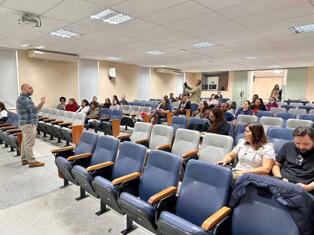 A imagem mostra um auditório ou sala de aula durante uma apresentação. Palestrante: No lado esquerdo, um homem de camisa xadrez azul e calça clara está em pé, gesticulando enquanto fala. Público: Várias pessoas estão sentadas nas poltronas, que têm estofado azul e detalhes em madeira nas laterais. O público parece estar acompanhando atentamente a apresentação. Ambiente: É um espaço amplo, com iluminação de teto embutida (painéis quadrados), paredes claras e poltronas dispostas em fileiras organizadas. Equipe: Há um cinegrafista em pé no meio da sala, registrando o evento com uma câmera. Parece ser um ambiente acadêmico ou corporativo.