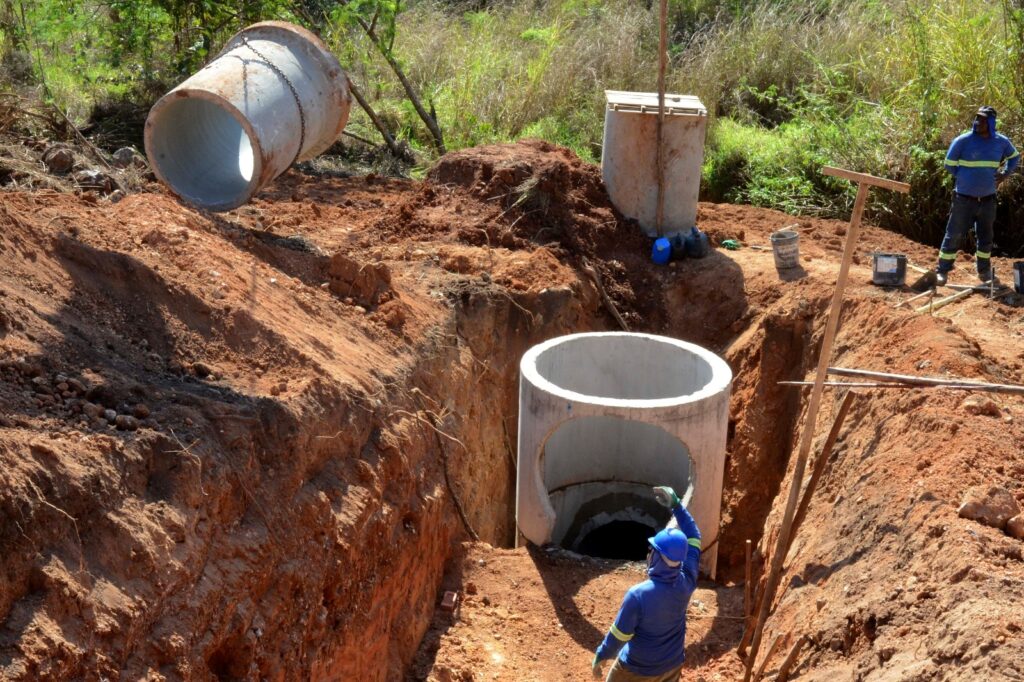 A cena mostra um canteiro de obras onde trabalhadores instalam grandes tubos de concreto em uma vala profunda escavada em solo avermelhado. Um dos segmentos já está posicionado no fundo da trincheira, enquanto outro, preso a uma corrente, aguarda para ser colocado. Dois operários com roupas de proteção e capacetes supervisionam o processo — um dentro da vala e outro na borda, garantindo precisão e segurança.