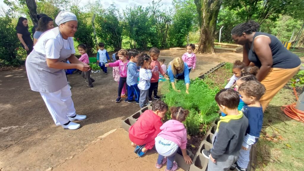 A imagem mostra um grupo de crianças participando de uma atividade de jardinagem em uma horta ao ar livre. Elas observam e ajudam a cuidar de um canteiro com plantas verdes, acompanhadas por duas adultas. À esquerda, uma profissional vestida de branco segura uma pequena muda ou planta. À direita, outra adulta orienta as crianças, que estão reunidas ao redor do canteiro e algumas se agacham para tocar a terra e as plantas. O espaço é arborizado, ensolarado e cercado por vegetação, com uma mangueira enrolada próxima à horta.