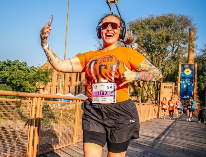 Uma corredora vibrante tira uma selfie enquanto participa de uma corrida 5K em uma ponte de madeira, celebrando seu progresso.
A imagem mostra uma mulher caucasiana sorridente, de cabelo loiro preso em um rabo de cavalo, usando óculos de sol laranja esportivos e fones de ouvido pretos, enquanto corre em uma ponte de madeira. Ela está vestindo uma camiseta laranja vibrante com um design gráfico e o número 1187 em um distintivo de corrida, acompanhado de "5K". Suas pernas estão vestidas com shorts de corrida pretos sobre leggings pretas. Braços e ombros da mulher exibem tatuagens coloridas e elaboradas, e ela tem tatuagens em seu braço esquerdo também. Ela está segurando um smartphone em sua mão esquerda, tirando uma selfie, e seu polegar direito está levantado em um gesto de "joinha" e o sinal de shaka com a mão esquerda. A ponte de madeira, com uma grade laranja, se estende para o fundo. Atrás dela, há uma fileira de outros corredores em suas próprias camisetas laranjas. Ao fundo, uma estrutura alta de tijolos vermelhos com uma chaminé, árvores verdes exuberantes e uma ponte decorada com lanternas azuis e um grande sol ou lua amarela que parecem ser parte de um evento ou decoração. A atmosfera é de alegria, energia e celebração, sugerindo um dia ensolarado e um evento de corrida de sucesso. O humor é edificante e inspirador.