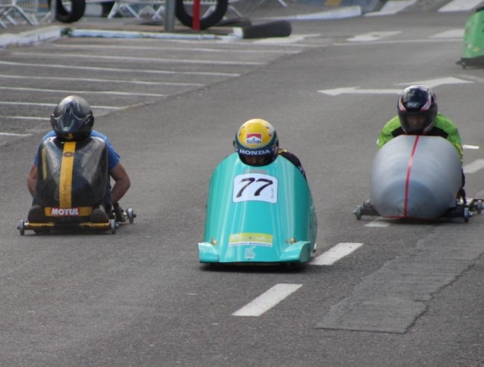 Na imagem há uma largada/trecho de corrida com três carrinhos de corrida sem motor (tipo soapbox/lowrider) alinhados na pista: Disposição: três competidores lado a lado, ocupando as faixas da rua. Carrinhos: o do centro é carenado, de cor verde-água, com o número 77 grande na dianteira; o da esquerda é mais simples, sem carenagem completa, com uma faixa vertical amarela no dorso e um adesivo “MOTUL” na traseira; o da direita tem carenagem cinza com uma linha vermelha central. Pilotos: todos em posição muito baixa/abaixada dentro dos carrinhos, usando capacetes integrais — o do centro tem capacete amarelo com adesivos (um deles com a palavra “HONDA” na viseira). Pista e ambiente: asfalto com faixas de sinalização branca; ao fundo notas de barreiras metálicas e pneus usados como proteções, sugerindo área vedada para a prova. Atmosfera: cena de competição organizada, com equipamentos de segurança e veículos compactos projetados para velocidade em descida.