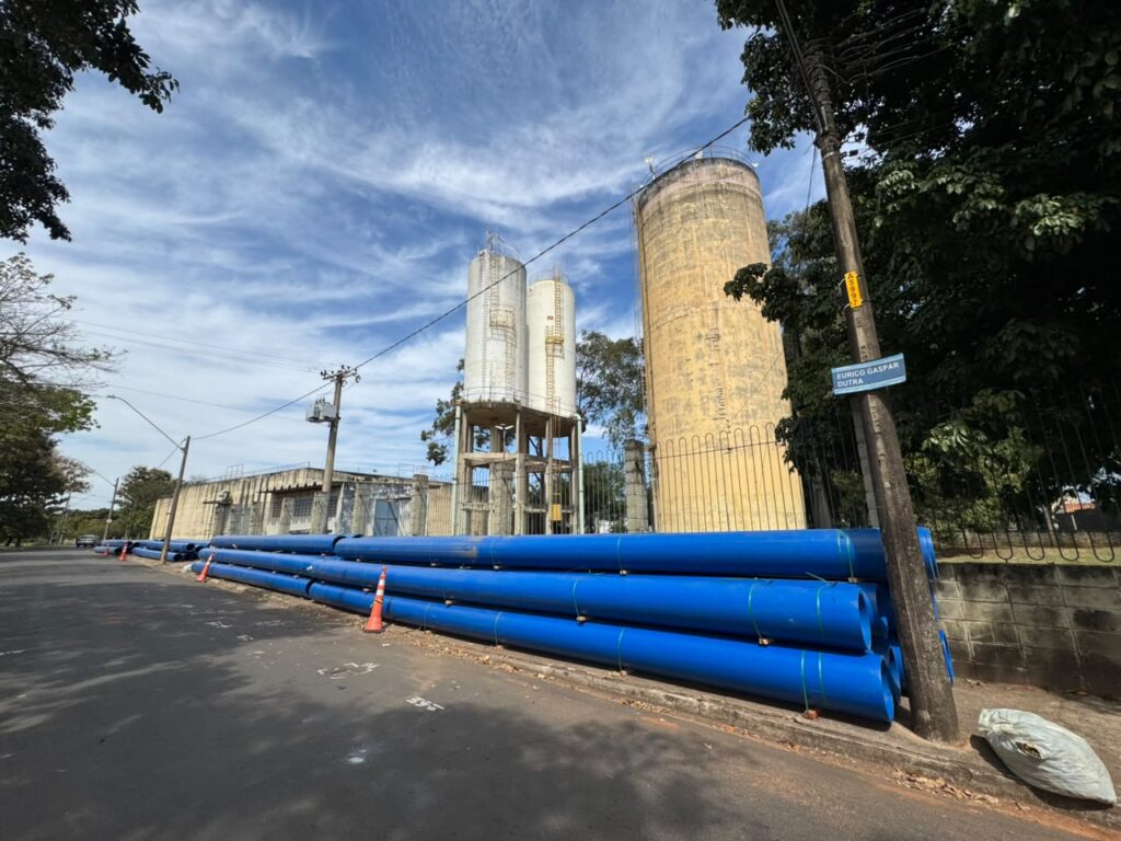 A imagem apresenta uma cena externa com uma perspectiva baixa e ampla, capturando uma rua ladeada por árvores e infraestrutura industrial. No primeiro plano, uma pilha generosa de tubos azuis grandes, com algumas amarrações verdes, está disposta ao longo da calçada. A superfície dos tubos é lisa e brilhante, refletindo a luz do sol. Três cones de trânsito laranja estão estrategicamente colocados entre os tubos, sugerindo um local de trabalho ou uma área restrita. Ao lado direito da imagem, próximo aos tubos, há um saco de lixo branco amassado e aberto. Dominando o fundo, encontram-se duas estruturas industriais de grande porte. À esquerda, há um edifício de concreto com uma aparência mais baixa e comprida, com janelas retangulares escuras e uma cobertura em telhado aparente. Centralizado na imagem e um pouco mais atrás, há um conjunto de dois silos cilíndricos metálicos, um ligeiramente mais alto que o outro, ambos com uma aparência envelhecida e com um padrão de degraus ou suportes visíveis em sua estrutura. À direita das duas torres menores, há uma torre cilíndrica muito maior, com uma cor amarelada distinta e uma textura que sugere concreto ou material similar. Esta torre maior tem uma base com uma série de arcos ou suportes verticais. Ao lado da torre amarela, há um poste de iluminação pública ou de fiação elétrica de madeira escura, com um sinal azul retangular fixado nele, onde se pode ler "EURICO GASPAR DUTRA". O poste está envolto por uma vegetação densa e escura, parte de uma árvore frondosa que se estende pela parte superior direita da imagem. Outras árvores com folhagem verde exuberante margeiam a cena, especialmente à esquerda e no fundo, com sombras projetadas no asfalto da rua. O céu é um elemento proeminente, apresentando um azul vibrante com nuvens brancas e finas, em forma de filamentos alongados, que criam um padrão interessante e dinâmico. Há postes e fios de eletricidade visíveis atravessando o céu, conectando-se à infraestrutura no solo. As cores predominantes na imagem são o azul intenso dos tubos, o amarelo desbotado da torre maior, o cinza e branco dos silos e do edifício, o verde das árvores e o azul claro do céu com o branco das nuvens. A composição é dominada pelas linhas horizontais dos tubos e da rua, contrastando com as linhas verticais das torres, do poste e dos fios. A perspectiva baixa, vista da rua, amplia a sensação de escala dos objetos, especialmente dos tubos e das estruturas industriais. A iluminação sugere um dia ensolarado, com sombras definidas no asfalto. Os detalhes nas estruturas, como o desgaste e os padrões, conferem um senso de realismo e antiguidade à cena, enquanto a limpeza e a organização dos tubos indicam uma atividade de construção ou manutenção em andamento.