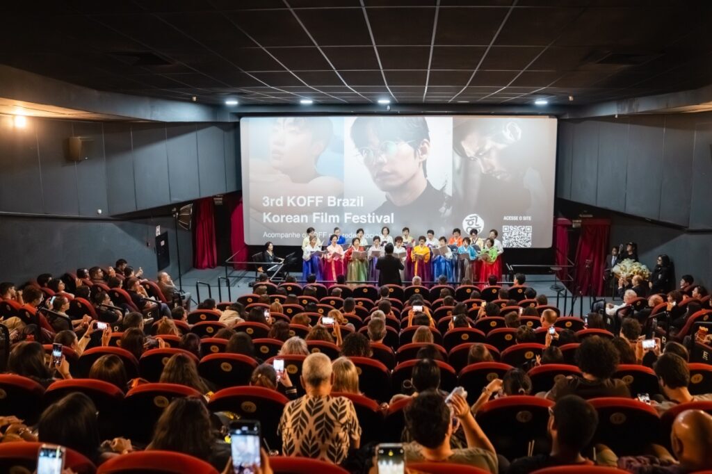 Esta imagem registra um evento cultural acontecendo dentro de uma sala de cinema. Aqui estão os detalhes principais: O Evento: Trata-se do "3rd KOFF Brazil - Korean Film Festival" (3º Festival de Cinema Coreano no Brasil). O nome do evento e algumas fotos de atores/artistas coreanos estão projetados em um telão grande atrás do palco. O Palco: Em frente à tela, há um coral formado por um grupo de mulheres vestindo hanboks coloridos (vestes tradicionais coreanas em tons de vermelho, azul, amarelo e verde). Elas estão segurando pastas e cantando, regidas por uma pessoa de costas para a plateia. Há também uma pianista acompanhando o grupo à esquerda do palco. A Plateia: A sala de cinema está cheia. O público está sentado nas poltronas vermelhas típicas de cinema e muitas pessoas estão segurando seus celulares no alto, gravando ou tirando fotos da apresentação. Ambiente: O ambiente é escuro, como é comum em salas de exibição, com a iluminação concentrada no palco e no telão. No canto superior direito da tela, há um código QR para acessar o site do evento.