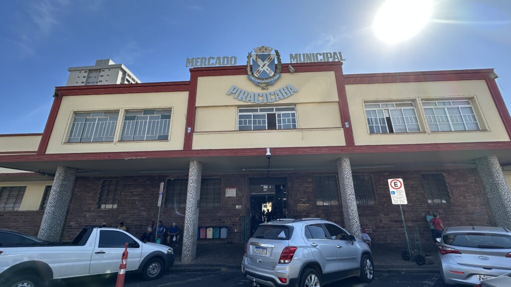 A imagem mostra a fachada do Mercado Municipal de Piracicaba, um edifício de estilo tradicional com colunas decoradas e paredes em tons de bege e vermelho. Acima da entrada principal, há um brasão com o nome da cidade, reforçando a identidade do espaço como ponto central da vida comunitária. Na frente do prédio, alguns veículos estão estacionados, incluindo uma caminhonete branca e um SUV prateado, enquanto pessoas circulam ou descansam próximas à entrada. Ao fundo, ergue-se um prédio mais alto, contrastando com a arquitetura histórica do mercado. O céu claro e ensolarado dá à cena um ar vibrante, ressaltando o papel do mercado como espaço de encontro e comércio local.