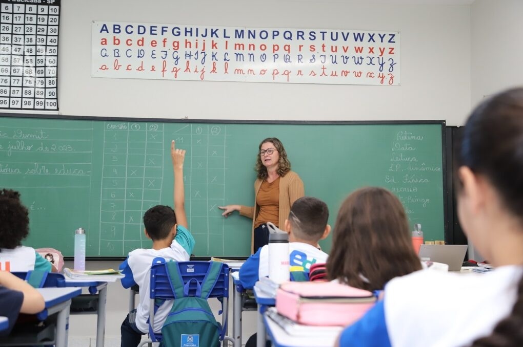 Esta é uma sala de aula dinâmica e organizada. Ação principal: Uma professora está em pé, à frente da lousa verde, apontando para uma tabela de marcações enquanto um aluno levanta a mão para interagir. Ambiente: A sala está bem equipada, com carteiras individuais, materiais escolares sobre as mesas (como garrafas de água e estojos) e mochilas penduradas nas cadeiras. Detalhes: Na parte superior da lousa, há um cartaz educativo com o alfabeto, apresentando letras em letra de forma e cursiva. A lousa contém uma tabela de "Questões" (1 e 2) com marcações de "x", além de uma lista de tópicos escritos à mão à direita (incluindo "Leitura", "Ed. Física", "Recreio", etc.). É uma cena clássica e acolhedora de aprendizagem ativa.