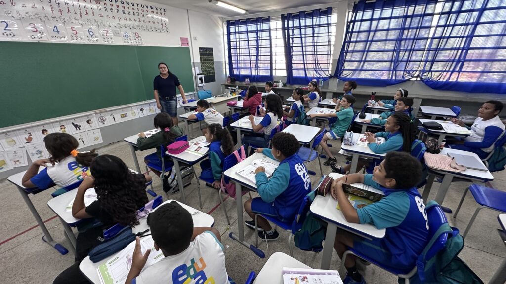 Nesta imagem, vemos uma sala de aula organizada, com alunos sentados em carteiras azuis, atentos à professora que conduz a lição diante de um grande quadro verde. Os estudantes, uniformizados em azul e branco, têm cadernos e livros abertos, mostrando envolvimento com a atividade. As paredes estão decoradas com materiais pedagógicos, como o alfabeto em letras maiúsculas e minúsculas, números acompanhados de ilustrações e desenhos que enriquecem o ambiente de aprendizagem