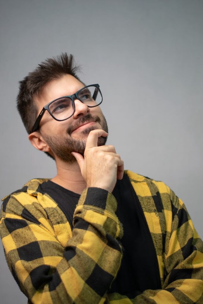 Esta é uma foto estilo retrato de um homem jovem, posando de forma pensativa e descontraída contra um fundo cinza neutro. Aqui estão os detalhes: O Homem: Ele tem cabelos escuros curtos, com um pouco de topete, e uma barba rala bem feita. Ele usa óculos de grau com armação preta quadrada. A Pose: Ele está com a mão no queixo, olhando para cima e para o lado com um sorriso leve e um olhar reflexivo, como se estivesse tendo uma ideia interessante ou imaginando algo. Vestimenta: Ele veste uma camisa xadrez de flanela com quadrados grandes em tons de amarelo e preto, aberta sobre uma camiseta preta básica por baixo. Estilo: A iluminação é suave e focada no rosto, o que confere à imagem uma qualidade profissional, parecida com uma foto de perfil ou de catálogo. O contraste da camisa xadrez amarela com o fundo neutro dá um destaque especial ao visual.