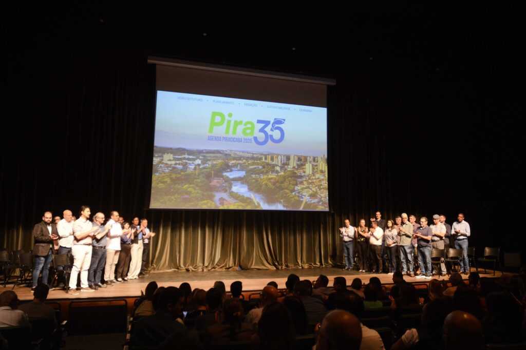 A imagem registra um momento coletivo no palco durante o evento de lançamento da Agenda Piracicaba 2035 – Pira 35. No centro superior, um grande telão exibe o logotipo “Pira 35”, acompanhado da frase “Agenda Piracicaba 2035” e de uma imagem panorâmica da cidade. Acima do logotipo aparecem os pilares Visão de Futuro, Planejamento, Inovação, Sustentabilidade e Cidadania. No palco, representantes da sociedade civil e do poder público estão reunidos em grupos, aplaudindo e celebrando o momento. Em primeiro plano, o público ocupa a plateia, observando a cerimônia. A fotografia transmite uma atmosfera de união, participação coletiva e compromisso com o planejamento do futuro de Piracicaba.