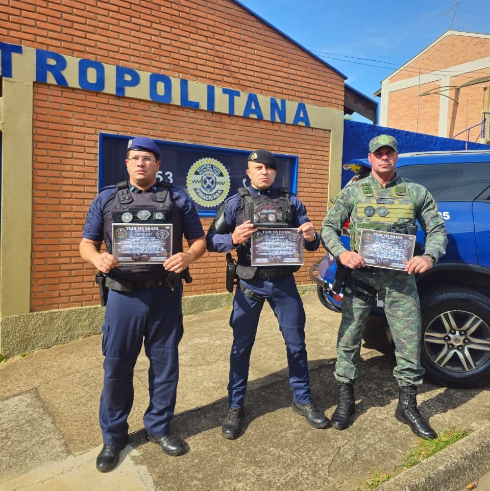 A imagem mostra três agentes de segurança uniformizados posando em frente a um prédio de tijolos com a inscrição “METROPOLITANA” parcialmente visível. Eles estão segurando certificados que parecem ser de reconhecimento ou conclusão de curso, com o título “TEAM SIX ELITE GYM” e o termo “CERTIFICADO” em destaque.