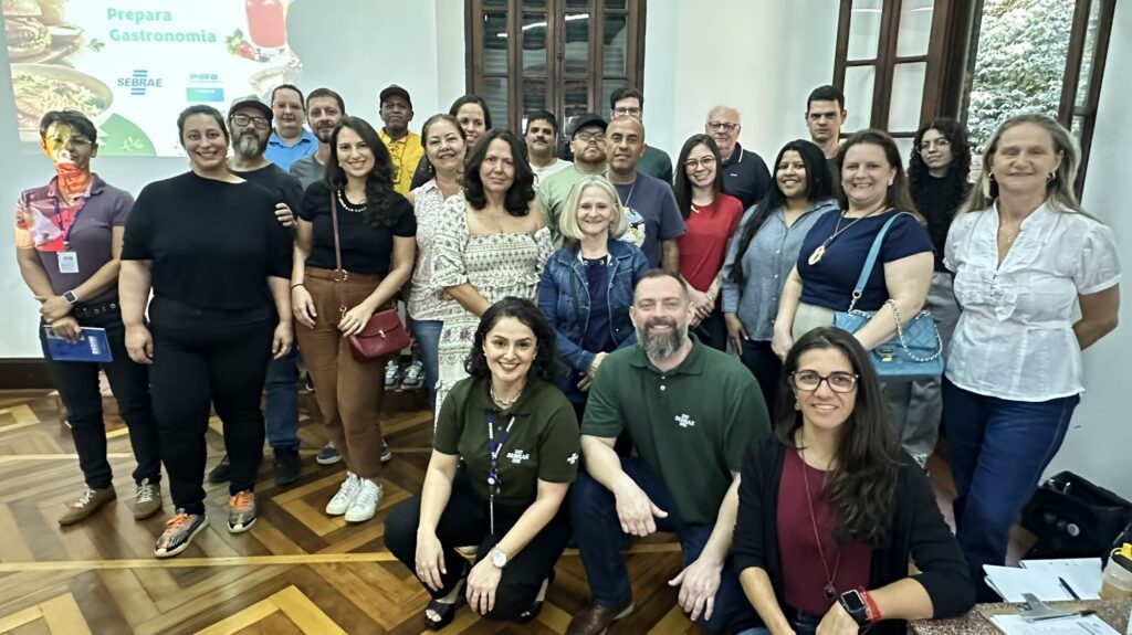 A imagem mostra um grupo de cerca de vinte pessoas reunidas em uma sala para uma foto coletiva. O grupo é composto por homens e mulheres de diversas idades, a maioria vestida de forma casual ou esporte fino. Três pessoas aparecem agachadas na frente (duas mulheres e um homem com barba de camisa polo verde), enquanto o restante está em pé logo atrás. Todos sorriem para a câmera. Ao fundo, no canto superior esquerdo, há uma tela de projeção onde se lê a frase "Prepara Gastronomia", acompanhada pelos logotipos do SEBRAE e da marca PIRA Turismo. O local possui janelas altas no estilo clássico com molduras de madeira e vidro. O piso é em parquet de madeira com padronagem em espinha de peixe/marquetaria clara e escura.
