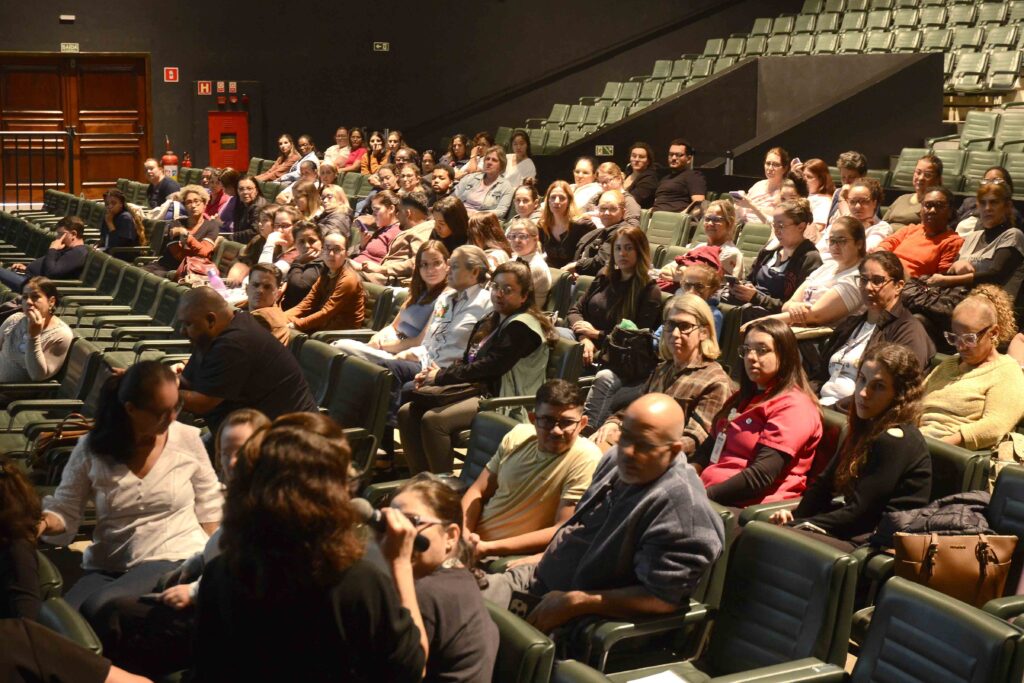 A imagem mostra um auditório lotado, com muitas pessoas sentadas em fileiras de poltronas verdes. O público parece assistir a uma apresentação ou evento, sob iluminação quente. À esquerda há portas de madeira e placas de saída de emergência. No fundo e nas laterais, veem-se corredores, corrimãos e fileiras superiores de assentos. A fotografia foi tirada de lado, destacando a grande quantidade de espectadores.