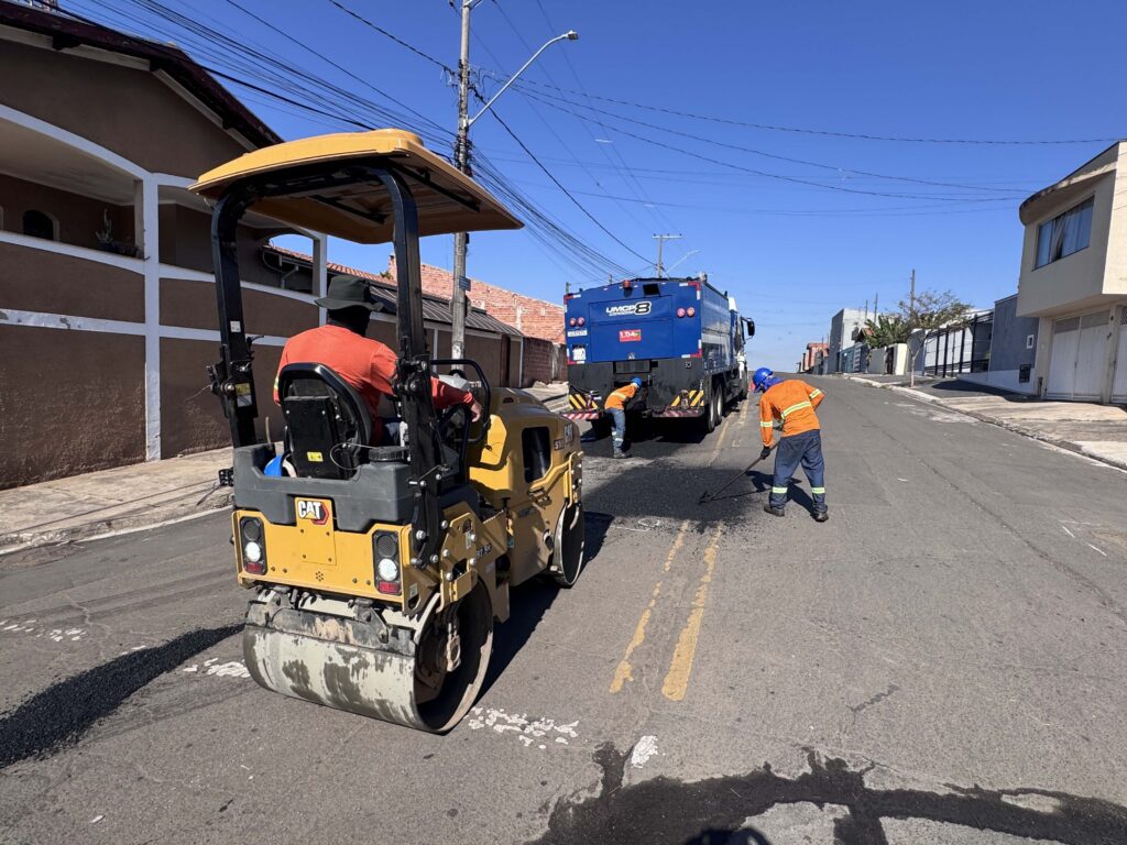 A imagem mostra uma equipe de trabalhadores usando uniformes laranja e azul realizando manutenção em uma rua residencial ensolarada. Em primeiro plano há uma máquina compactadora amarela da Caterpillar; ao fundo, um caminhão azul da empresa “UMCP 8”. As casas têm fachadas simples, e o céu está limpo, com muitos fios elétricos presos aos postes.