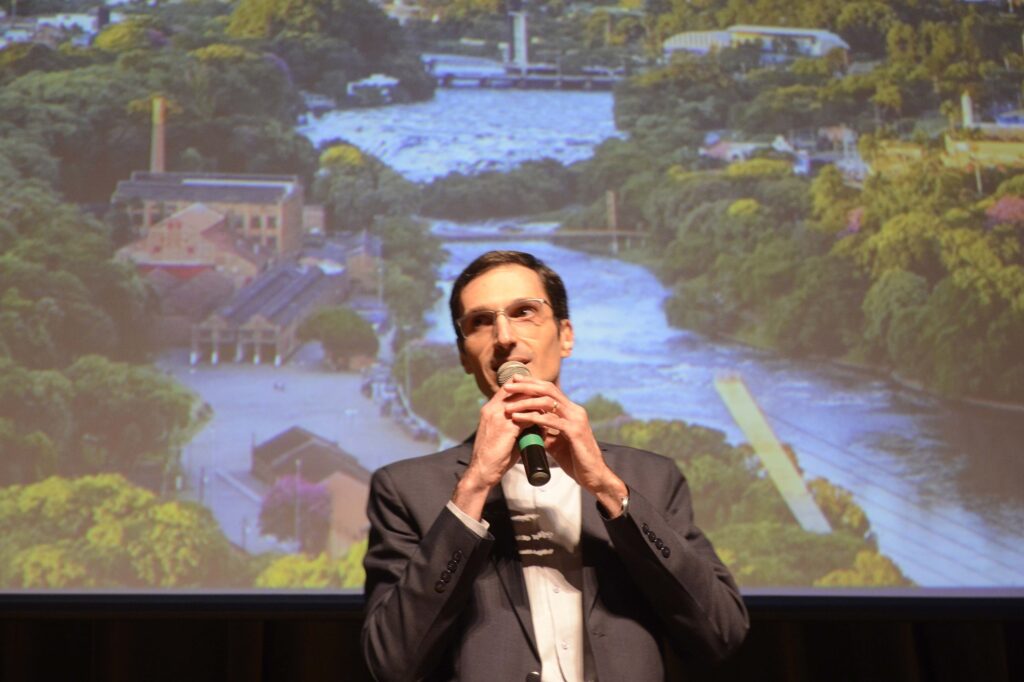 Uma fotografia captura um homem de meia-idade falando ao microfone durante uma apresentação, com uma imagem projetada de uma paisagem urbana com um rio ao fundo, destacando um momento profissional. A composição da cena é centrada no indivíduo, que ocupa a maior parte do plano médio inferior, criando um contraste direto entre sua figura em primeiro plano e o cenário projetado atrás dele. Ele segura o microfone com ambas as mãos próximas ao rosto, focando a atenção do espectador na sua expressão e no ato de comunicação. A imagem de fundo serve como um pano de fundo visual que contextualiza o ambiente da palestra ou evento, criando uma sobreposição entre o orador e o local representado na projeção. O sujeito principal é um homem de aparência madura, com cabelos curtos escuros, óculos de grau com armação clara e um rosto de traços finos. Ele mantém uma expressão concentrada e serena enquanto fala. Ele está vestido com uma camisa branca sob um terno cinza escuro, exibindo um visual formal e sóbrio. A postura é ereta e compenetrada, com suas mãos posicionadas de forma a segurar o microfone com firmeza, mas com elegância, demonstrando que está habituado a falar em público. A imagem é uma fotografia digital que documenta um evento real, apresentando características técnicas típicas de registros de conferências. A execução prioriza o foco nítido no orador, enquanto o fundo projetado possui uma profundidade de campo ligeiramente mais suave. O estilo é jornalístico ou institucional, visando registrar a interação entre o palestrante e o conteúdo visual exposto em uma tela grande, mantendo o realismo das cores e a iluminação característica de espaços internos com projeções. O ambiente é um auditório ou sala de conferências com iluminação controlada para permitir a visualização da projeção ao fundo. A luz que incide sobre o homem vem de fontes frontais ou laterais, iluminando seu rosto e destacando a textura de seu terno, enquanto a projeção ao fundo ilumina a parte posterior da cena com uma luminosidade fria, proveniente da imagem de um rio e edificações em um dia claro. A atmosfera geral é de seriedade e engajamento intelectual, típica de ambientes onde ocorre a disseminação de informações, apresentações de projetos ou palestras educativas.