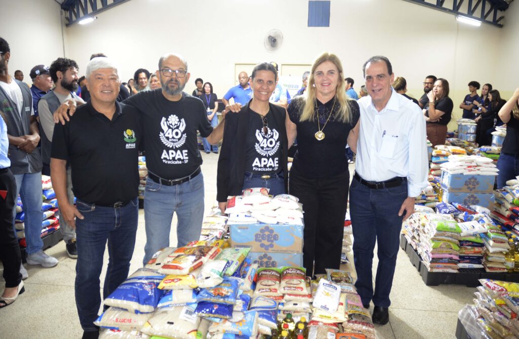 A imagem mostra um grupo de cinco pessoas sorrindo e posando juntas em um ambiente interno, cercadas por pilhas de alimentos não perecíveis — como arroz, feijão, açúcar e óleo — organizados em pacotes e caixas. Duas pessoas vestem camisetas pretas com o logotipo da APAE Piracicaba - SP, comemorando seus 40 anos, o que indica um evento solidário ou de celebração institucional. Ao fundo, há mais participantes observando e interagindo, reforçando o clima de união e engajamento comunitário.