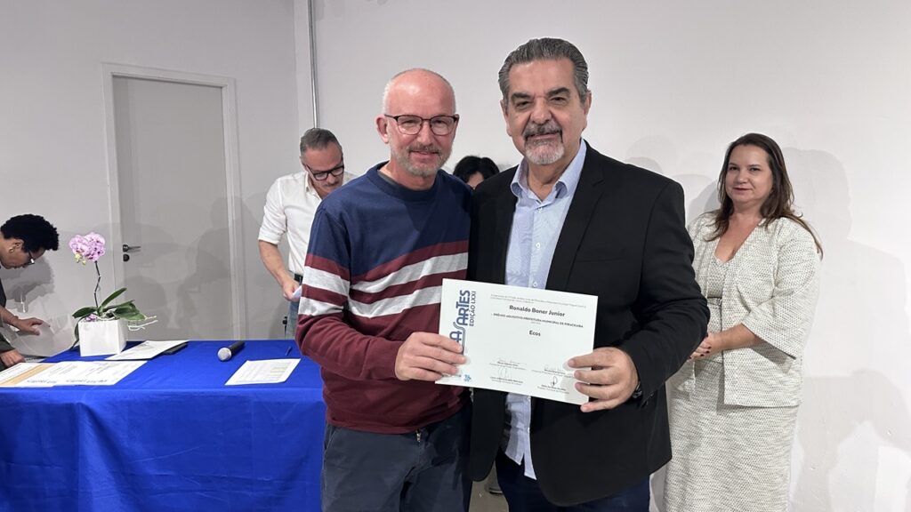 A foto captura um momento de premiação em um evento cultural. Em primeiro plano: Dois homens estão lado a lado posando para uma foto. O da esquerda é um homem de óculos e barba, vestindo um suéter com faixas horizontais nas cores bordô, azul marinho e cinza. Ele segura um certificado da "Bela Artes - Edição LXXI". O homem à direita é mais velho, com barba grisalha, vestindo uma camisa social clara por baixo de um blazer preto. O Certificado: O documento premiado é entregue a "Ronaldo Boner Junior", com o título da obra "Ecos", recebendo o "Prêmio Aquisitivo Prefeitura Municipal de Piracicaba". Cenário: Eles estão em frente a uma mesa coberta com uma toalha azul, onde há um vaso com uma orquídea rosa e alguns papéis. Ao fundo, à esquerda, um homem de camisa branca está ocupado com documentos sobre a mesa. À direita, uma mulher de blusa bege (um conjunto texturizado) observa a cena, sorrindo levemente. O fundo é uma parede branca lisa e neutra.