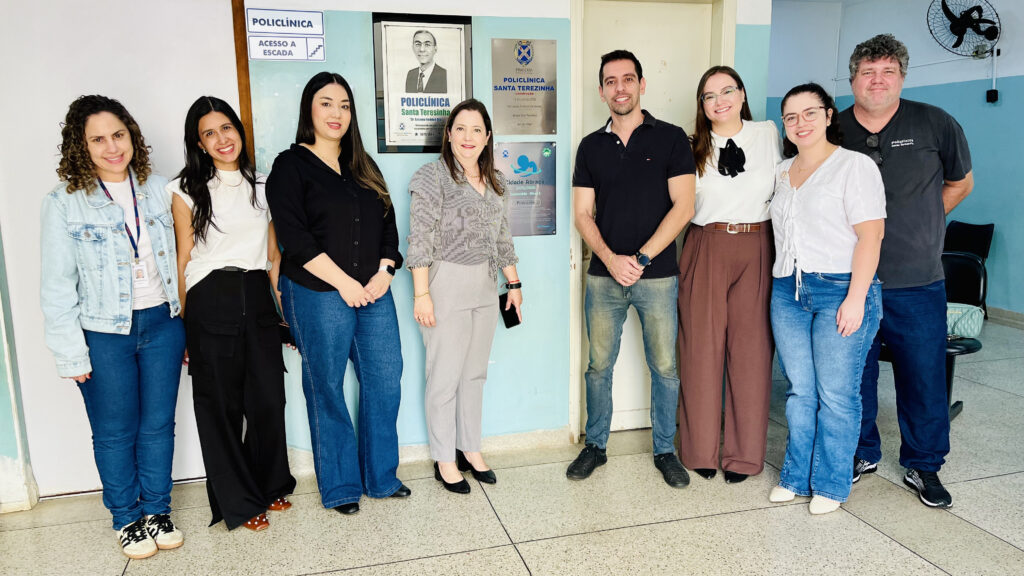Uma fotografia em grupo registra oito pessoas posando sorridentes em um ambiente interno de uma policlínica, transmitindo uma atmosfera de colaboração profissional e harmonia entre os indivíduos presentes no local. A cena é composta por uma fileira de oito adultos posicionados lado a lado, ocupando o centro da imagem em uma formação horizontal frente a uma parede institucional. O enquadramento é do tipo plano geral, permitindo capturar o corpo inteiro dos participantes e o cenário de fundo, que inclui placas comemorativas e sinalização da instituição. A disposição dos integrantes cria uma composição equilibrada, onde as posturas eretas e os olhares voltados para a câmera sugerem um momento de registro formal, possivelmente após um evento ou reunião de trabalho, mantendo uma interação harmoniosa e estática. Os oito sujeitos apresentam estilos variados, equilibrando o traje casual com o profissional. As quatro mulheres à esquerda vestem combinações de calças jeans ou sociais com blusas ou jaquetas leves, exibindo cabelos em diferentes estilos, desde lisos até cacheados. À direita, os três homens e a mulher que os acompanha mantêm o mesmo padrão de informalidade elegante; os homens optam por camisas polo e jeans, enquanto a mulher usa uma blusa branca de tecido leve e calça de alfaiataria em tons terrosos. Todos demonstram expressões faciais gentis e sorridentes, mantendo posturas naturais e relaxadas, com as mãos posicionadas ao longo do corpo ou entrelaçadas à frente. A imagem é uma fotografia digital capturada sob condições de iluminação ambiente natural, que banha o cenário de forma uniforme, evitando sombras intensas e mantendo a nitidez dos detalhes. O estilo é documental e institucional, focado na clareza das fisionomias e na identificação do ambiente de fundo. A execução técnica privilegia o foco frontal, garantindo que todos os integrantes estejam bem iluminados e com boa resolução, sendo uma escolha fotográfica prática para registros de equipes profissionais, priorizando a visibilidade e o contexto informativo em vez de artifícios estilísticos elaborados. O cenário é o interior de uma policlínica, caracterizado por paredes de tons claros, com azulejos azul-claros e brancos que conferem um aspecto clínico e limpo. Ao fundo, elementos como placas de identificação e um quadro com uma foto ilustram a localização e a importância do espaço. O piso é de material claro e neutro, complementando a claridade do ambiente. A iluminação é difusa e clara, típica de espaços internos com luz artificial, criando uma atmosfera serena e acolhedora. O ambiente de trabalho institucional é reforçado pela disposição organizada e pelo comportamento profissional do grupo, que reflete coesão e um sentimento de orgulho pelo local de atuação.