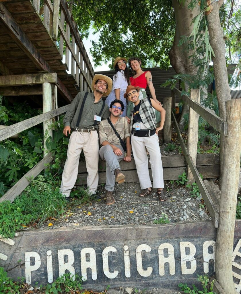 Esta imagem mostra um grupo de cinco jovens posando para uma foto em uma escadaria de madeira em um ambiente ao ar livre, possivelmente um ponto turístico ou parque em Piracicaba. Aqui estão os detalhes observados: O Grupo: Cinco pessoas sorridentes, vestidas com trajes casuais e rústicos (alguns usam chapéus e suspensórios), sugerindo que podem estar participando de uma atividade cultural, teatral ou apenas aproveitando um passeio temático. O Cenário: Eles estão em uma escadaria de madeira rústica, cercada por vegetação e árvores, com uma placa de madeira grande e rústica em primeiro plano onde se lê claramente "PIRACICABA" pintado com letras brancas. Detalhes: À esquerda, um rapaz usa chapéu, camisa listrada, suspensórios e calças claras. No centro, um rapaz sentado usa óculos, camisa social bege e calça cinza. À direita, outro rapaz usa chapéu, colete xadrez, camisa preta e calça clara. Atrás, duas moças completam o grupo: uma usando um chapéu de palha e camisa branca, e a outra de blusa vermelha. Atmosfera: O clima é descontraído, de amizade e celebração, com boa iluminação natural de dia ensolarado.