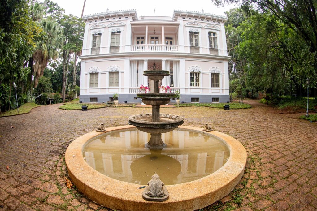 Esta imagem apresenta um belo casarão histórico, muito bem conservado, com uma arquitetura clássica e elegante. Aqui estão os detalhes observados: O Casarão: É um edifício de dois andares com uma fachada pintada em tons suaves de rosa e branco. Possui diversas janelas altas com molduras detalhadas, colunas na entrada principal e uma varanda no andar superior com uma balaustrada branca. O estilo remete a construções coloniais ou neoclássicas. O Jardim/Pátio: Em frente ao casarão, há um pátio pavimentado com pedras irregulares. No centro, destaca-se uma fonte circular de pedra, com vários níveis, que parece conter água parada. Detalhes da Fonte: A fonte possui pequenas esculturas de sapos decorando a borda, o que adiciona um toque charmoso e peculiar ao conjunto. Entorno: O casarão está cercado por uma área arborizada com árvores altas e densas, o que confere um ar de serenidade e isolamento. A iluminação sugere um dia nublado ou luz suave, destacando a textura da arquitetura e o verde ao redor.