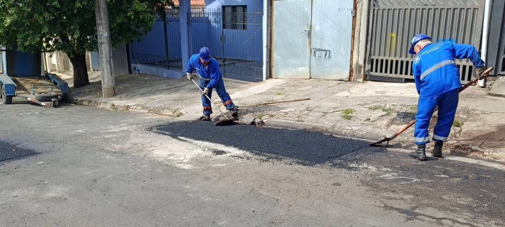 Na imagem, dois trabalhadores uniformizados de azul e usando capacetes de segurança realizam a manutenção de uma rua em um bairro residencial. Eles espalham e nivelam o asfalto fresco com pás e rastelos, cobrindo um trecho da via próximo à calçada. Ao fundo, aparecem casas com portões e uma árvore que oferece sombra, compondo um cenário cotidiano de infraestrutura urbana, onde o trabalho de reparo garante melhores condições de tráfego e segurança para a comunidade.