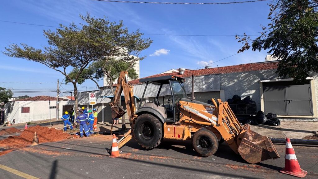 A cena ocorre em uma área urbana sob um céu azul claro com nuvens esparsas. Uma escavadeira retroescavadora laranja está estacionada na rua, com seu braço dianteiro abaixado. A máquina, com a marca "CASE" visível em seu corpo, tem pneus grandes e uma cabine de metal. Uma grande pilha de terra avermelhada está ao lado da escavadeira, com alguns cones laranja e brancos posicionados estrategicamente perto dela e da máquina. Um grupo de trabalhadores em uniformes azuis e amarelos, vestindo capacetes amarelos, está reunido perto da escavadeira, parecendo supervisionar ou participar do trabalho. Atrás deles, à direita, há um edifício com telhado de telha vermelha e paredes claras, com uma entrada grande e metálica. Sacos pretos de lixo estão empilhados em frente a esta entrada. À esquerda, há uma casa branca com janelas escuras e algumas árvores com folhagem verde densa ao redor. Cabos elétricos correm horizontalmente no céu. A rua em primeiro plano tem marcas amarelas no asfalto. A composição é dominada pela escavadeira laranja e pela terra avermelhada, com os trabalhadores e os edifícios formando o cenário. A iluminação sugere um dia ensolarado.