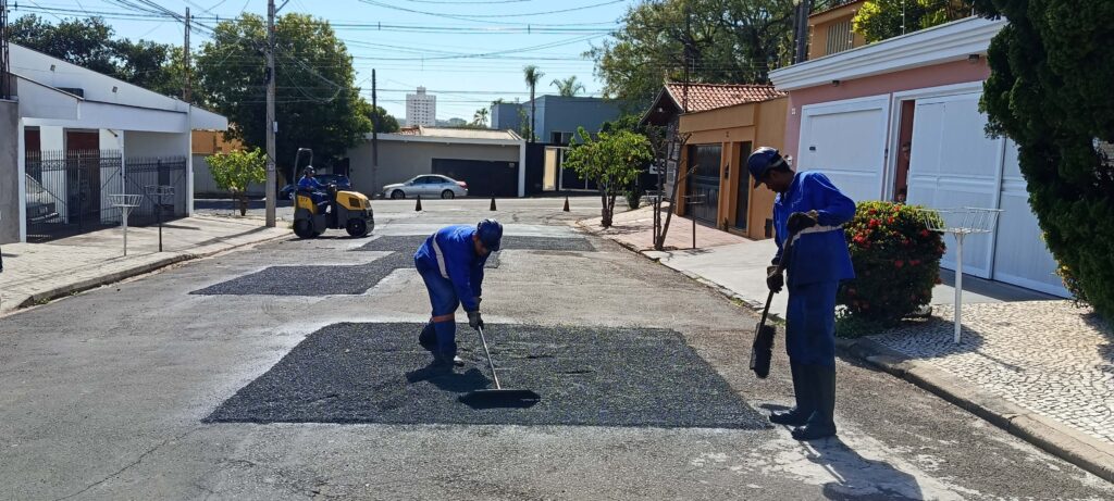 dois trabalhadores com uniformes azuis e capacetes realizam a manutenção de uma rua residencial. Eles espalham o asfalto em remendos retangulares da pista utilizando ferramentas manuais, enquanto ao fundo outro profissional opera um pequeno rolo compressor amarelo para compactar o material. O cenário mostra casas alinhadas, árvores e postes com fiação aérea, compondo um ambiente urbano típico.