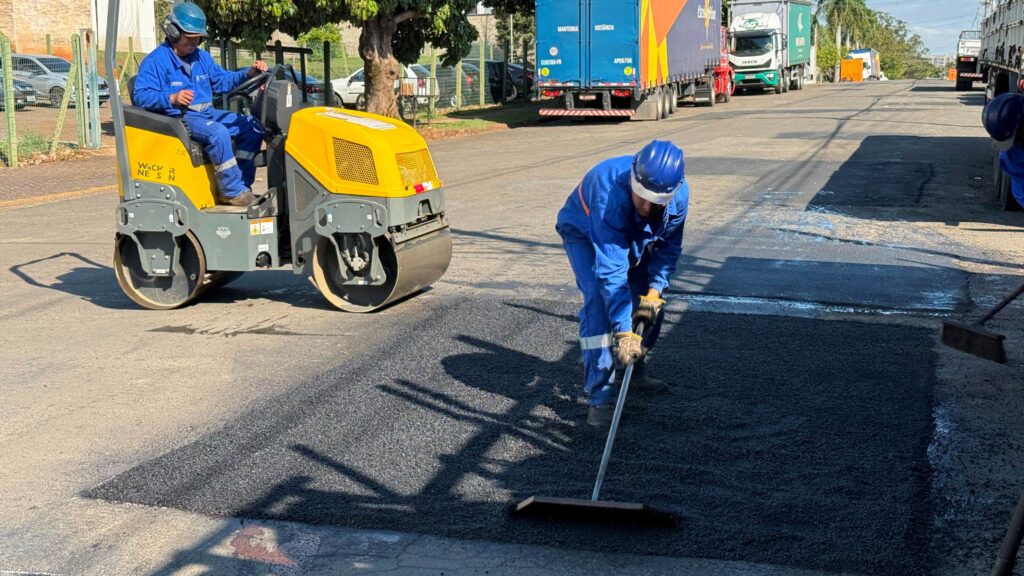 Na cena, dois trabalhadores com uniformes azuis e capacetes da mesma cor atuam na recuperação de um trecho de pavimento: um deles manobra um rolo compactador amarelo, que prensa e alisa o asfalto recém-aplicado, enquanto o outro espalha e nivela o material escuro com uma ferramenta semelhante a um ancinho; ao fundo, aparecem caminhões estacionados, uma árvore e uma área cercada.
