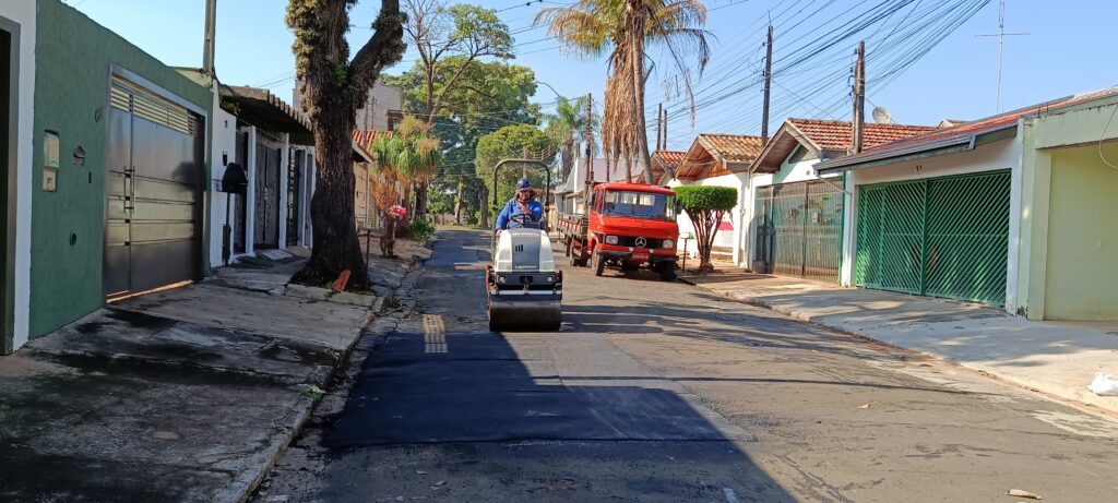 Na imagem vemos uma rua residencial durante um trabalho de pavimentação: Em primeiro plano, um rolo compactador pequeno (vibro-compactador) está avançando sobre uma faixa de asfalto fresco recém-aplicada. Um operário conduz a máquina; ele usa capacete e roupa de trabalho azul. À direita, estacionado na rua, há um caminhão vermelho (marca Mercedes) e, ao fundo, mais adiante, outro trecho da via com árvores e casas. As calçadas são de concreto; de um lado há garagens com portões verdes e, do outro, muros e portas de casas. Há várias árvores ao longo da rua, inclusive uma palmeira alta; postes com muitos cabos elétricos cruzam o céu. Céu limpo e iluminação solar forte, sombras bem definidas, indicando que a foto foi tirada em dia claro.